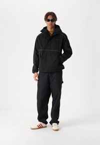 Black pullover jacket with a hood, front pocket, and reflective stitching, paired with dark pants and sneakers with red stripes.