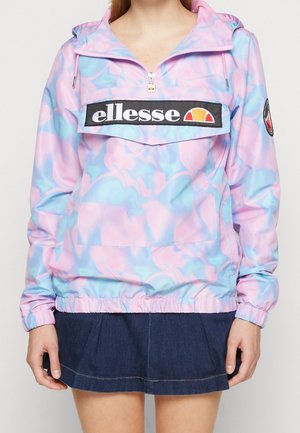 Kobieta ubrana w pastelową różowo-niebieską bluzę typu pullover z czarną naszywką z logo „ellesse” oraz ciemną spódnicę dżinsową.