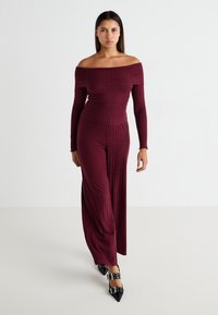 Burgundy bordázott overál, vállpánt nélküli nyakkivágással és testhez simuló felsőrésszel; széles lábú nadrág; fekete, hegyes orrú magassarkúval párosítva.