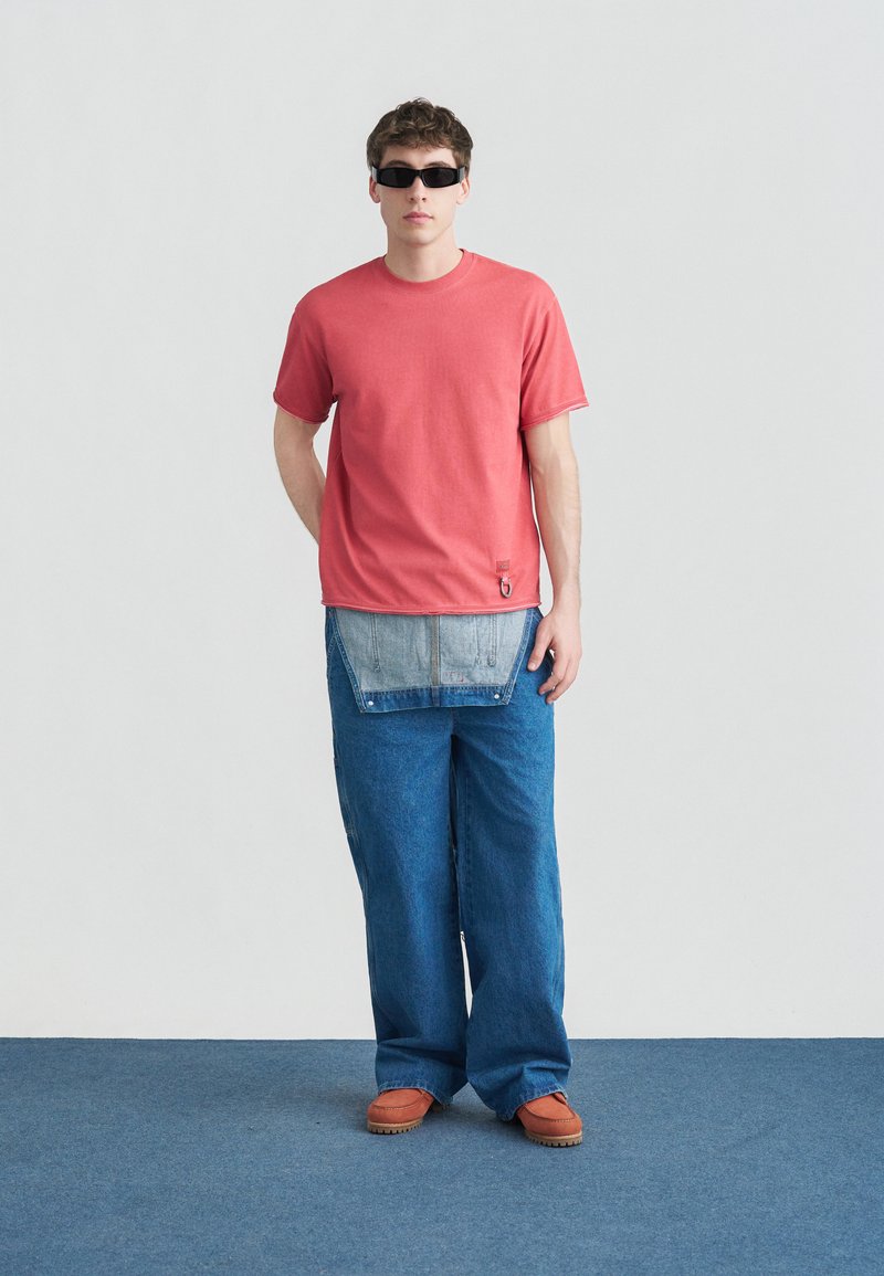 Giovane uomo con occhiali da sole neri, t-shirt corallo, jeans larghi blu con grembiule di denim attaccato e scarpe marroni, in piedi su un tappeto blu.