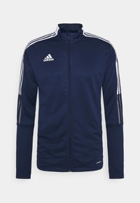 Chaqueta deportiva de color azul marino con rayas blancas en los hombros y las mangas, cuello alto, cremallera frontal completa y logo de "adidas" en la parte superior izquierda del pecho.