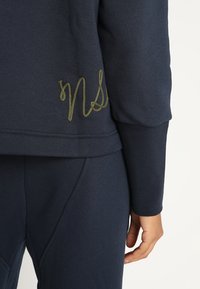 Acercamiento de una persona que viste una sudadera y pantalones azul marino, mostrando un bordado en letras doradas "nS" en la parte baja de la espalda de la sudadera.