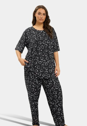 Femme portant un haut ample noir et un pantalon avec des motifs de cœurs blancs et roses, debout, une main dans la poche, sur un fond uni.