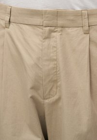 Pantalon en coton beige avec passants pour ceinture, une fermeture éclair à l’avant et un design plissé sur la ceinture avant.