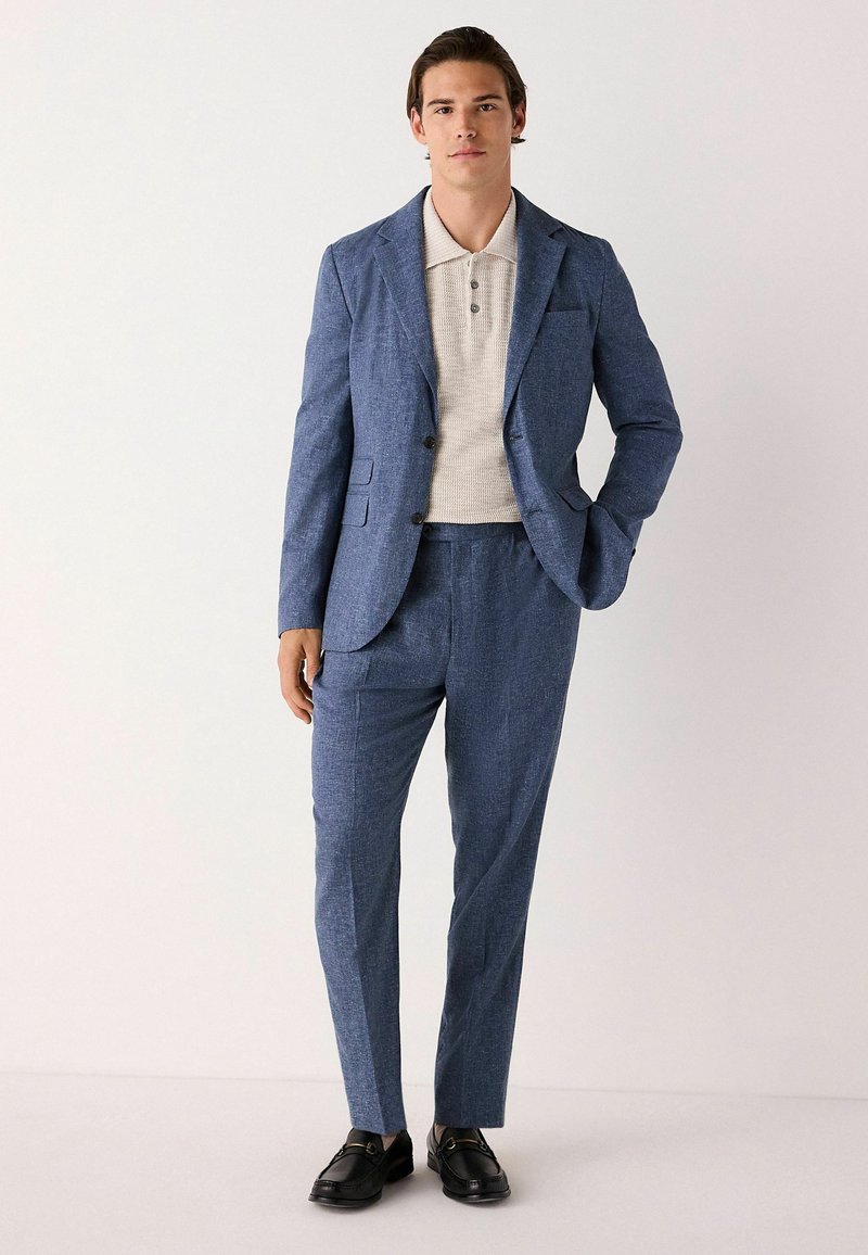 Blau strukturierte Anzugjacke und passende Hose kombiniert mit einem hellbeigen Poloshirt. Schwarze Dressschuhe. Minimalistisches Design, taillierte Silhouette.