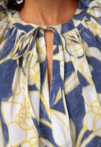 Pusero, jossa on kukkakuosi ja sinivalkoinen väriteema keltaisilla yksityiskohdilla. Pehmeä kangas, kerätty kaula-aukko ja solmukuvio.