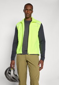High-visibility yellow vest with reflective accents, lightweight fabric, zip closure, worn over a dark long-sleeve shirt, held with a gray helmet.