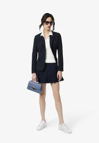 Blazer marine sur un top en tricot blanc, associé à une jupe plissée marine. Tient un sac à main bleu et porte des baskets blanches ainsi que des lunettes de soleil élégantes.