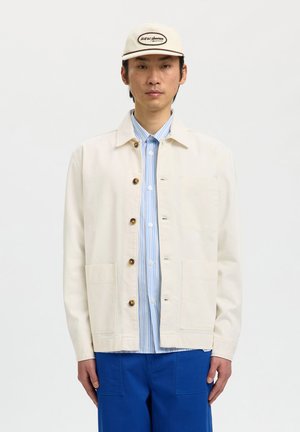 Mężczyzna ubrany w kremową zapinaną na guziki kurtkę, niebieską koszulę w paski, niebieskie spodnie oraz beżową czapkę z logo na białym tle.