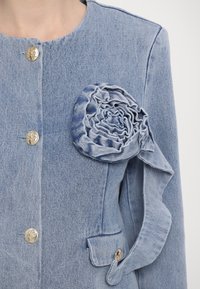 Veste en jean bleu clair avec des boutons dorés, un grand détail en rosace en tissu sur la poitrine et une poche à rabat avec un bouton doré.