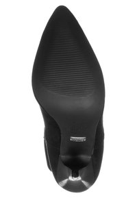 Scarpa nera in suede con tacco alto, punta affusolata, suola testurizzata con righe orizzontali, design con apertura alla caviglia e accento nero lucido vicino al tacco.