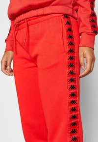 Person wearing bright red sweatpants with black logo tape down side seams and matching red drawstring waistband.