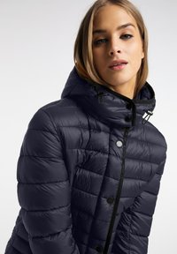 Marineblauwe gewatteerde jas met capuchon, met een gequiltde textuur, contrasterende zwarte hardware en een rits- en knoopsluiting aan de voorkant.