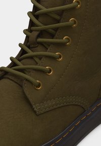 Gros plan d'une botte en cuir vert foncé avec des lacets assortis, des œillets métalliques dorés, des coutures détaillées et une semelle noire avec des coutures jaunes.