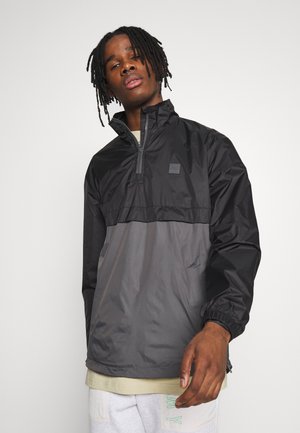 Sort og grå letvægts zippered pullover jakke med en halv længde for-zipper, elastiske manchetter og et lille logo patch på brystet.
