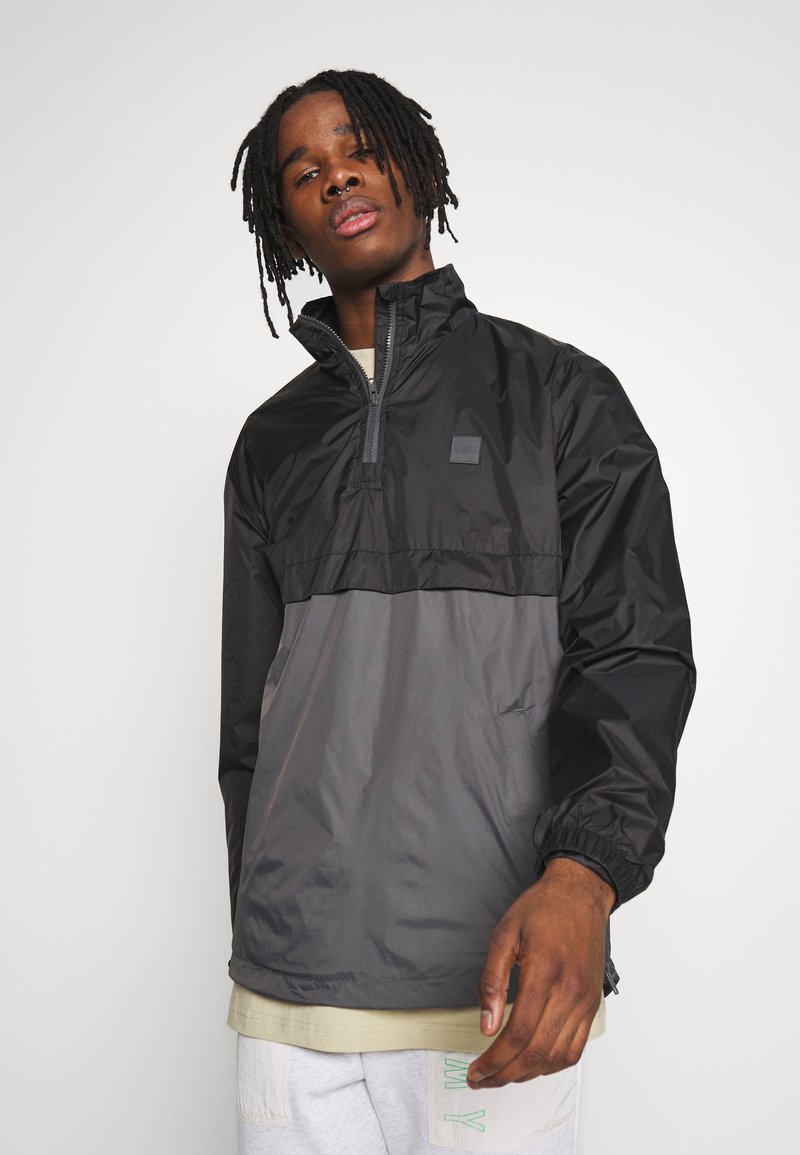 Sort og grå letvægts zippered pullover jakke med en halv længde for-zipper, elastiske manchetter og et lille logo patch på brystet.