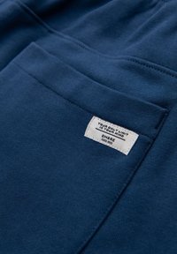 Close-up van blauwe katoenen stof met een zak waarop een wit label staat met de tekst "JOUW ENIGE BEPERKING IS JE GEEST" en de merknaam "OMBRE."