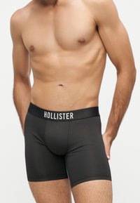 Μυώδες ανδρικό κορμί που φοράει μαύρα boxer της Hollister με ελαστική μέση και εμφανές όνομα μάρκας, στέκεται με το ένα χέρι στο πλάι.