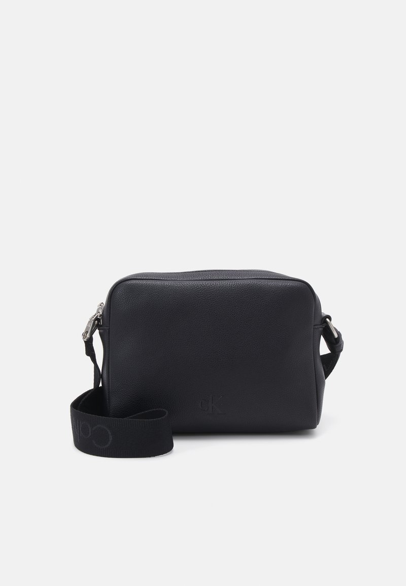 Bolso cruzado de cuero negro con forma rectangular, cierre de cremallera y una correa de tela con un logo. Superficie texturizada con un marcado sutil.