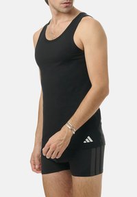 Black athletic tank top made of smooth fabric, featuring a round neckline and logo accent. Worn with black mesh shorts.