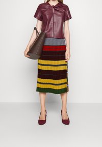 Top in pelle bordeaux con dettaglio a nodo, abbinato a una gonna midi plissettata a righe multicolori in verde, giallo e rosso. Borsa marrone e scarpe intrecciate bordeaux.