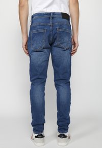 Jeans de denim azul de corte ceñido, con una cinturilla estándar, dos bolsillos traseros y una textura suave, combinados con zapatillas blancas y grises.