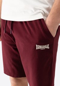 Bordeaux shorts lavet af blødt bomuld, med en snoretræk i taljen, sidelommer og en hvid logo tekst på venstre lår.