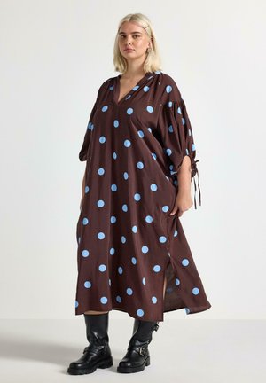 Donna in piedi che indossa un vestito largo marrone con grandi pois blu, maniche legate a metà braccio e stivali neri fino a metà polpaccio.