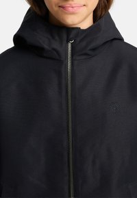 Giacca nera con cappuccio e zip frontale, caratterizzata da una texture liscia e un piccolo logo sul lato sinistro. Design semplice e moderno.
