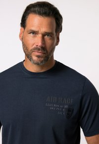 Námořnická modrá tričko vyrobené z bavlny, s kulatým výstřihem a předním designem s embosovaným textem a logem umístěným na levé straně.