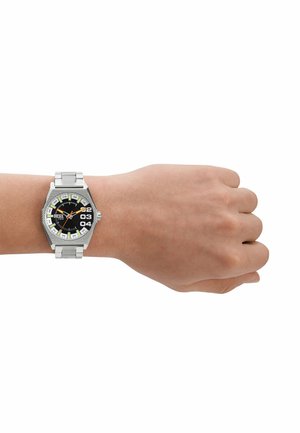 Montre en métal argenté avec un cadran hexagonal, un fond noir, des index horaires verts et blancs, et des accents oranges sur les aiguilles, présentée sur une main.