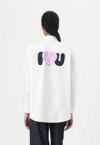 Weißes langärmliges Shirt mit abgerundetem Saum. Auf der Rückseite befindet sich ein großes schwarzes und lila grafisches "I ❤️ U" mit "HGO" in der Mitte. Aus Baumwollmaterial.