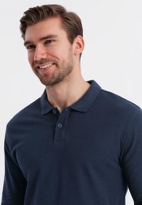 Leende man med kort brunt hår och skägg som bär en mörkblå långärmad polo mot en enfärgad vit bakgrund.