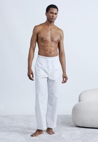 Λευκό βαμβακερό παντελόνι lounge με ελαστική μέση. Απλός σχεδιασμός με άνετη εφαρμογή, φορεμένο ξυπόλυτο, τοποθετημένο σε ανοιχτό φόντο.