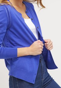 Modrý pletený cardigan s kulatým výstřihem, zapínáním na knoflíky a dlouhými rukávy. Má volný střih a žebrované manžety. Nošený přes bílý top.