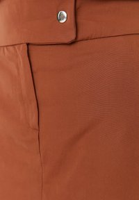 Tissu couleur rouille avec une poche avant et un bouton-pression argenté sur un vêtement, montrant les détails de la texture et des coutures.