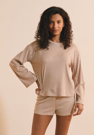 Donna con i capelli ricci indossa un top beige a righe a maniche lunghe e shorts coordinati, in piedi con una mano sul fianco contro uno sfondo beige semplice.