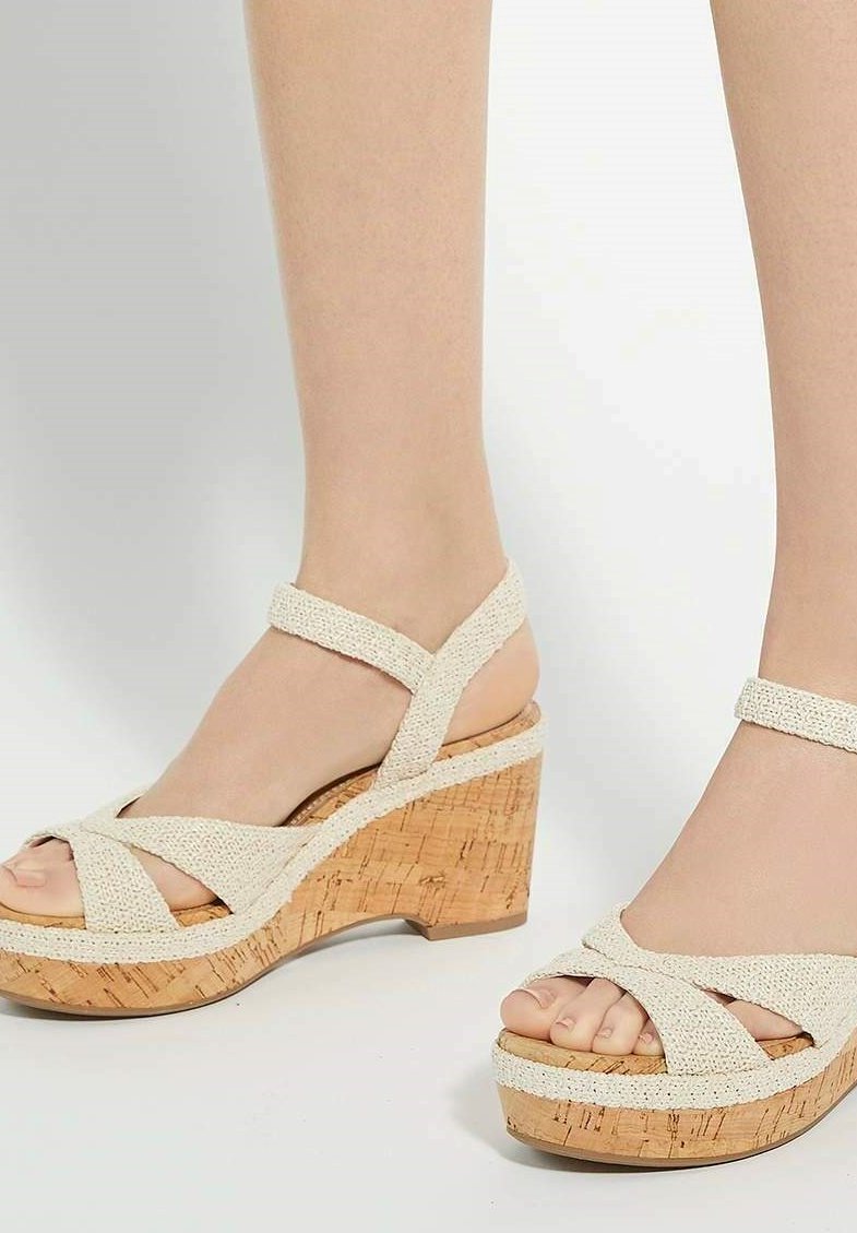 Wedge sandals featuring a textured beige fabric upper, crisscross strap design, and a cork-like platform sole. Open-toe style.