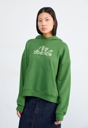 Un mannequin se tient devant un fond clair, portant un sweat à capuche vert avec "new balance" imprimé dessus, associé à une jupe en jean foncé.