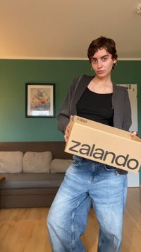 Kartonska škatla s tiskanimi besedami "zalando" v debelih črnih črkah. Oseba nosi črn zgornji del in široke, izbledele modre kavbojke. Zelen stena v ozadju.