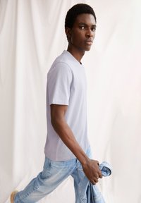 Helles blaues T-Shirt mit kurzen Ärmeln aus weichem Stoff, kombiniert mit hellblauen Jeans. Das Model hält in einer Hand eine gefaltete Jeansjacke.