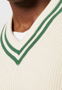 Gestrickter Pullover in Creme mit strukturiertem Muster, tiefem V-Ausschnitt und dunkelgrünen Streifen am Kragen.