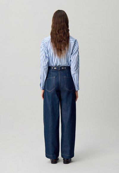 Jeans de mezclilla oscura de cintura alta con piernas anchas, cinco bolsillos y acentos de hardware plateado. Combinados con una camisa de botones a rayas azul y blanca.