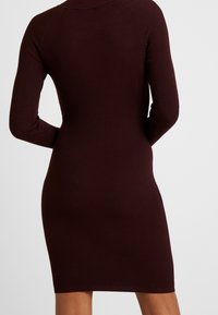 Robe ajustée à côtes de couleur bordeaux sombre, avec des manches longues et un col rond ; texture lisse dans un style moulant.