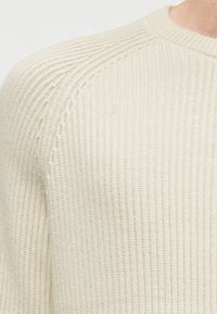 Pull en maille couleur crème avec texture côtelée, doté d'un col rond et de détails distinctifs aux coutures des épaules. Matière douce et légère.