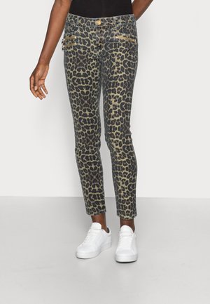 Pantalon slim avec un motif léopard foncé, détails de fermeture éclair dorée sur les poches avant, porté avec des baskets blanches et un haut noir.