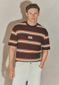 Maroonstribet t-shirt med orange, hvide og gule striber, med et centreret "PROVIDE" logo. Bæres med lyse jeans.
