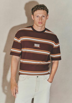 Maroonstribet t-shirt med orange, hvide og gule striber, med et centreret "PROVIDE" logo. Bæres med lyse jeans.