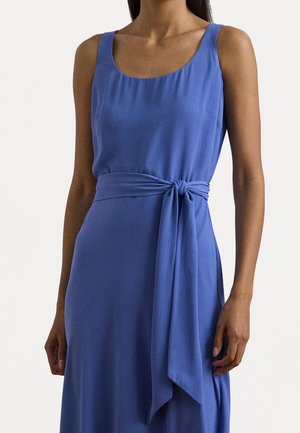 Mouwloze blauwe jurk met ronde hals en brede stoffen riem die in een knoop in de taille is gestrikt, gedragen door een staand persoon.