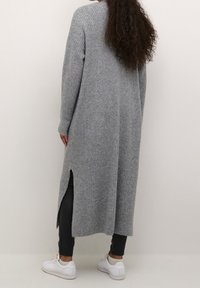 Long gray ribbed cardigan with side slits, soft texture, and fitted sleeves, paired with black leggings and white sneakers.